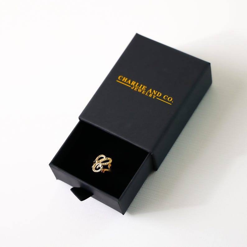 May include: A gold ring with a delicate, intricate design is displayed in a black box with the text "CHARLIE AND CO. JEWELRY" printed in gold.