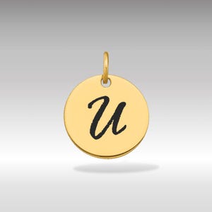 Pode incluir: Um pingente redondo dourado com a letra cursiva "U" preta no centro. O pingente tem uma pequena argola na parte superior para uma corrente. O fundo é um gradiente cinza claro.