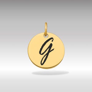 Pode incluir: Um pingente circular dourado com a letra cursiva "G" preta no centro. O pingente tem uma pequena argola na parte superior para prender a uma corrente ou pulseira. O fundo é um gradiente de cinza claro.
