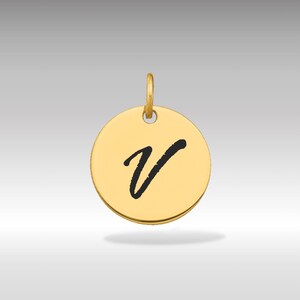 Pode incluir: Pingente circular dourado com a letra cursiva preta "V" no centro. O pingente tem uma pequena argola na parte superior para uma corrente. O fundo é um gradiente de cinza claro a branco.