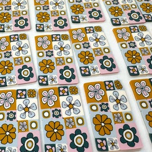 Op de afbeelding: Een verzameling handgemaakte keramische tegels met een retro bloemenpatroon. De tegels hebben een patchwork-ontwerp met roze, blauwe, witte, groene en gele bloemen op een witte achtergrond.