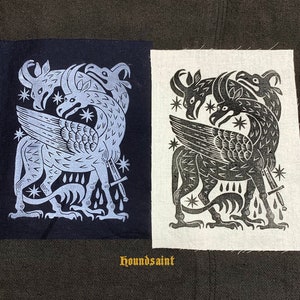 Puede incluir: Dos parches de tela con un diseño de criatura mítica. Uno es azul marino con una impresión azul claro, el otro blanco con una impresión negra. El diseño presenta una bestia alada con tres cabezas, una espada y acentos de estrellas. El texto "houndsaint" está en la parte inferior.
