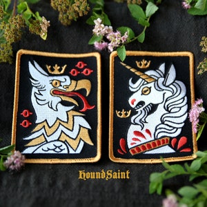Op de afbeelding: Twee geborduurde patches met zwarte achtergronden en gouden randen. Eén toont een witte griffioen met een gouden kroon en rode accenten. De andere toont een witte eenhoorn met een gouden kroon en rode accenten. De tekst "HoundSaint" staat onderaan.