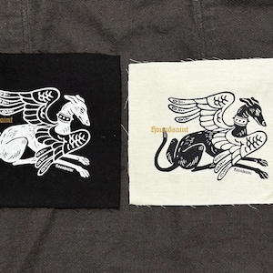 Op de afbeelding: Twee vierkante patches met een gevleugeld windhondontwerp. Eén patch is zwart met een wit ontwerp, de andere is gebroken wit met een zwart ontwerp. De tekst "houndsaint" staat boven de windhond.