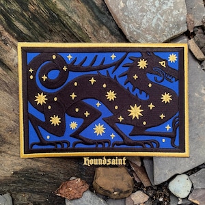 Peut inclure: Écusson brodé représentant un dragon noir orné d'étoiles dorées sur fond bleu. Le patch a une bordure dorée et le mot "houndsaint" dans une police stylisée.