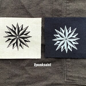 Op de afbeelding: Twee vierkante stoffen patches. Eén is gebroken wit met een zwart sterrenpatroon. De andere is marineblauw met een wit sterrenpatroon. Het woord "houndsaint" is in witte letters eronder gedrukt.