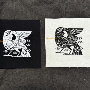 Op de afbeelding: Twee vierkante patches met een gestileerd vogelontwerp en meerdere ogen. Eén patch is zwart met een wit ontwerp, de andere is wit met een zwart ontwerp. De tekst "houndsaint" is in goud op beide patches gedrukt.