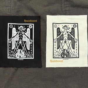 Op de afbeelding: Twee stoffen patches. Eén is zwart, de andere gebroken wit. Elk heeft een witte illustratie van een gevleugelde, hondachtige figuur met uitgestrekte armen en een slang. Het woord "houndsaint" staat op elk gedrukt.