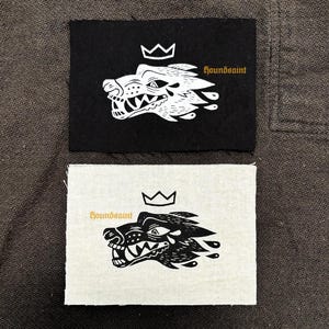 Op de afbeelding: Twee rechthoekige patches, één zwart en één gebroken wit, elk met een gestileerde wolfskop met een kroon en het woord "houndsaint". De wolf is wit, met een gedetailleerd ontwerp. De tekst is in een schreefletter.