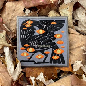 Könnte beinhalten: Quadratischer Aufnäher mit einem schwarzen Raben-Design, mehreren orangefarbenen Augen und dem Text "houndsaint" in Weiß. Der Aufnäher befindet sich auf einem grauen Hintergrund mit braunem Rand, umgeben von Herbstlaub.