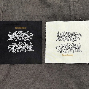 Op de afbeelding: Twee vierkante patches, één zwart en één gebroken wit, elk met een witte botanische illustratie van twee takjes met bessen. Het woord "houndsaint" is in goud op beide patches gedrukt.