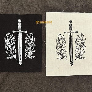 Op de afbeelding: Twee vierkante patches met een zwaard- en bloemenontwerp. Eén patch is zwart met een wit ontwerp, de andere is gebroken wit met een zwart ontwerp. De tekst "houndsaint" is gedrukt op de gebroken witte patch.