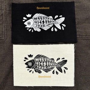 Op de afbeelding: Twee patches, één zwart en één gebroken wit, elk met een gedetailleerde witte illustratie van een vis met een kroon. De tekst "houndsaint" is in goud op elke patch gedrukt. Het visontwerp bevat waterdruppels.