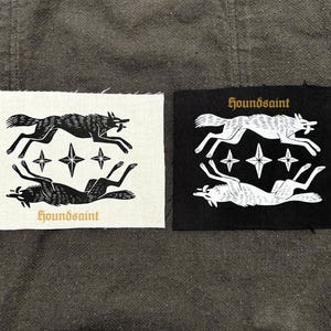 Op de afbeelding: Twee patches met een wolf ontwerp. Eén patch is wit met een zwart wolf ontwerp, sterren en het woord "houndsaint" in goud. De andere patch is zwart met een wit wolf ontwerp, sterren en het woord "houndsaint" in goud.