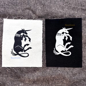 Può includere: Due toppe in tessuto rettangolari con un grafico di unicorno. Una toppa è bianca con un unicorno nero e il testo "houndsaint" in azzurro. L'altra toppa è nera con un unicorno bianco e il testo "houndsaint" in color oro.