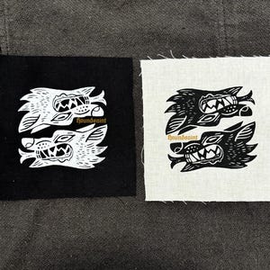 Op de afbeelding: Twee vierkante patches, één zwart en één gebroken wit, elk met een symmetrisch wolfontwerp in contrasterende kleuren. De tekst "houndsaint" is in goud onder de wolfskoppen gedrukt. De patches hebben een rafelige rand.