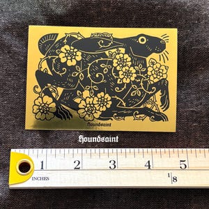 May include: A gold sticker with a black illustration of a rabbit surrounded by flowers and vines. The sticker is approximately 3 inches wide. The text "houndsaint" is printed in a stylized font at the bottom of the sticker.