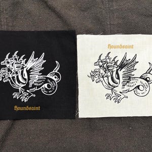 Op de afbeelding: Twee vierkante patches, één zwart en één gebroken wit, elk met een witte illustratie van een driekoppige draak met vleugels en een slangachtige staart. De tekst "houndsaint" is in goud op elke patch gedrukt.