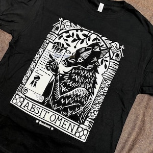 May include: A black t-shirt featuring a white graphic print of a wolf-like creature holding a quill and a scroll. The design is framed by an architectural archway with the text "ABSIT OMEN" and "houndsaint" printed at the bottom of the illustration.