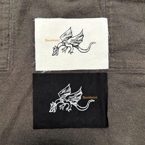 Op de afbeelding: Twee rechthoekige patches, één witte en één zwarte, elk met een gedetailleerde drakenillustratie in contrasterende kleuren. De tekst "houndsaint" is in een gestileerd lettertype op elke patch gedrukt, wat een unieke touch aan het ontwerp toevoegt.