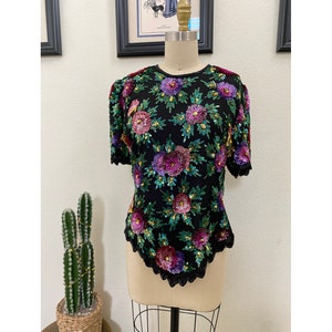 May include: A black sequined top with a floral design in shades of pink, purple, green, and gold. The top has short sleeves and a scalloped hem.