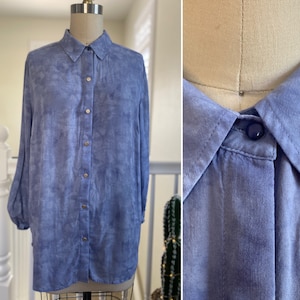May include: A long-sleeved, light blue button-down shirt with a tie-dye pattern. The shirt has a classic collar and a button closure.