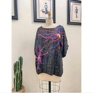 May include: A black sequined top with a colorful floral design. The design features pink, blue, and gold sequins. The top has short sleeves and a round neckline.