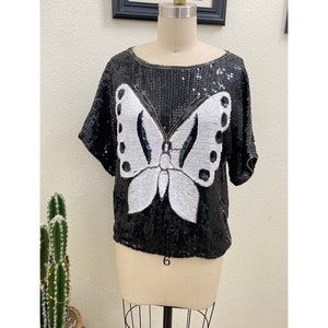 May include: A black sequined top with a white butterfly design. The top has short sleeves and a round neckline.