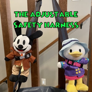 May include: The Adjustable Safety Harness is a blue and gray adjustable strap with a black buckle. The harness is hanging from a brown wooden railing. Two plush toys, one a black and white Mickey Mouse character and the other a yellow duck character, are attached to the harness. The text "THE ADJUSTABLE SAFETY HARNESS" is written in green letters on the top of the image. The text "LISTING INCLUDES ONE HARNESS. NUIMOS NOT INCLUDED." is written in white letters on the bottom of the image.