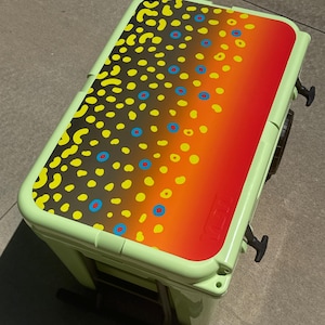 May include: A light green cooler with a lid featuring a colorful design. The lid has a gradient of orange to red, with yellow and blue dot patterns. The cooler has black handles and a black pull-out handle.