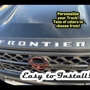 May include: A black truck with a silver "FRONTIER" lettering on the hood. The truck has a black grille with a red Nissan logo. The text "Personalize your Truck! Tons of colors to choose from!" and "Easy to Install!" are visible in the image.