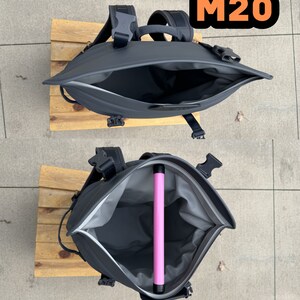 Puede incluir: Una mochila negra con una abertura con cremallera, que muestra el interior de la mochila. La mochila tiene un tubo rosa en el interior. La mochila tiene el texto "M20" en la parte superior.