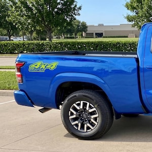 May include: A vibrant blue pickup truck with black and silver wheels. The truck features a "4x4 Frontier" decal in green and yellow. The vehicle is parked on a paved surface with green foliage in the background.