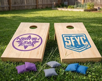 May include: Two wooden cornhole boards with a natural finish, each featuring a different logo in purple and blue. The boards are set up on a grassy lawn, with bean bags in matching colors scattered around.
