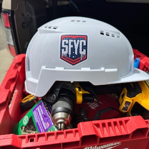 May include: A white hard hat with the letters "SFYC" on a sticker, resting in a red Milwaukee toolbox. The toolbox contains a drill, screws, and other tools. The hard hat is a safety item for construction or industrial work.