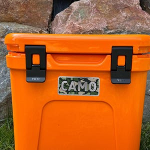 May include: Bright orange cooler with black latches and the word "YETI" on the latches. A "CAMO" sticker is on the front. The cooler is on green grass with a stone background.