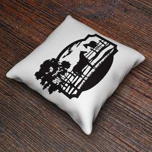 May include: White square throw pillow with a black silhouette design of a horse, fence, tree, and landscape. The pillow is on a dark wood surface.