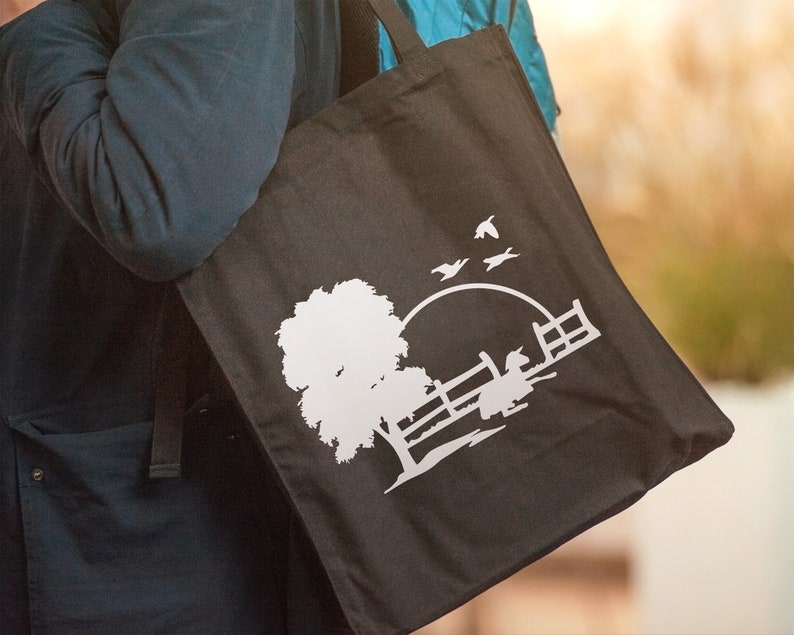 Peut inclure: Un sac fourre-tout noir avec un motif en silhouette blanche d'un arbre, une cl&ocirc;ture et des oiseaux volant au-dessus d'un soleil couchant.