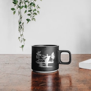 May include: Black ceramic mug with a white silhouette of a deer in a forest scene. The mug has a handle and is sitting on a wooden table.