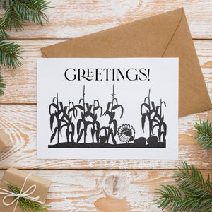 Op de afbeelding: Een witte kaart met de tekst "GREETINGS!" in het zwart gedrukt. De kaart toont een zwart silhouet van een kalkoen die in een ma&iuml;sveld staat.