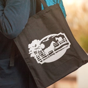 May include: Black tote bag featuring a white graphic of a horse, fence, and tree within a circular design. The bag is carried over a person's shoulder. The design evokes a rural or equestrian theme.