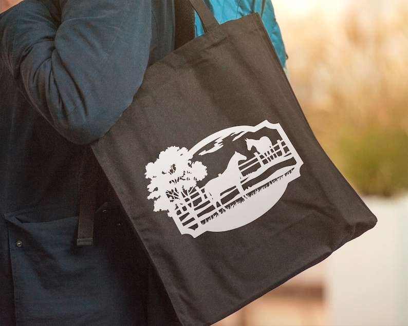 May include: Black tote bag featuring a white graphic of a horse, fence, and tree within a circular design. The bag is carried over a person's shoulder. The design evokes a rural or equestrian theme.