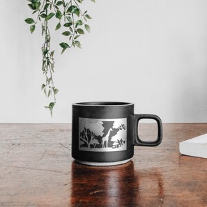 May include: Black ceramic mug with a white silhouette of a giraffe and trees. The mug has a handle on the right side.
