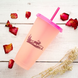 May include: A pink reusable cup with a pink straw and a silhouette of a deer in a forest scene. The cup is on a white wooden surface with dried rose petals scattered around it.