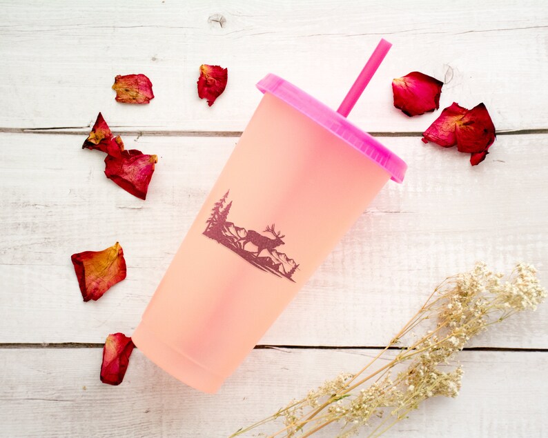 May include: A pink reusable cup with a pink straw and a silhouette of a deer in a forest scene. The cup is on a white wooden surface with dried rose petals scattered around it.