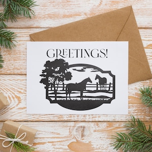 May include: A white greeting card with the word "GREETINGS!" in black script. The card features a black silhouette of two horses in a pasture with a tree. The card is on a wooden surface with a brown envelope, gifts, and pine branches.