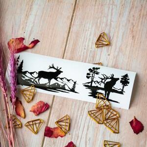 May include: Two black silhouette stickers of a deer in a forest setting. The stickers are on a white background and are surrounded by gold paper clips.