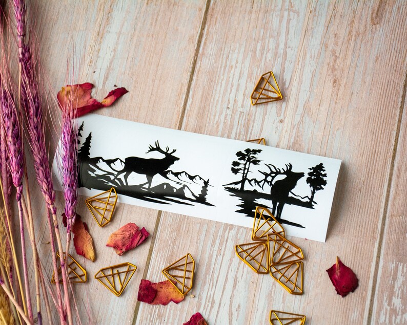 May include: Two black silhouette stickers of a deer in a forest setting. The stickers are on a white background and are surrounded by gold paper clips.