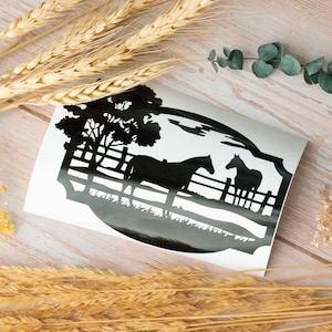 May include: A black and white decal featuring two horses in a pasture with a fence and trees. The design is set against a white background, with a silhouette style. The decal is on a light wood surface, with wheat stalks and greenery.