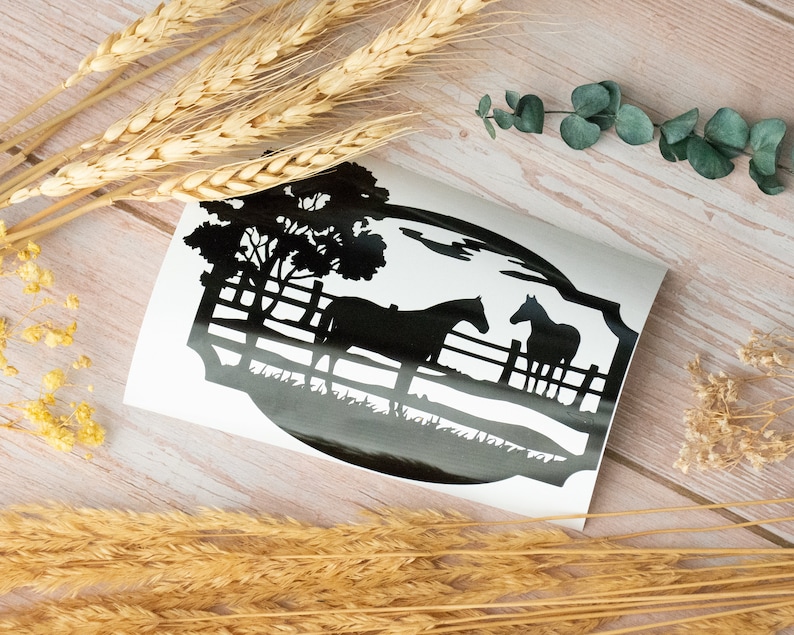 May include: A black and white decal featuring two horses in a pasture with a fence and trees. The design is set against a white background, with a silhouette style. The decal is on a light wood surface, with wheat stalks and greenery.